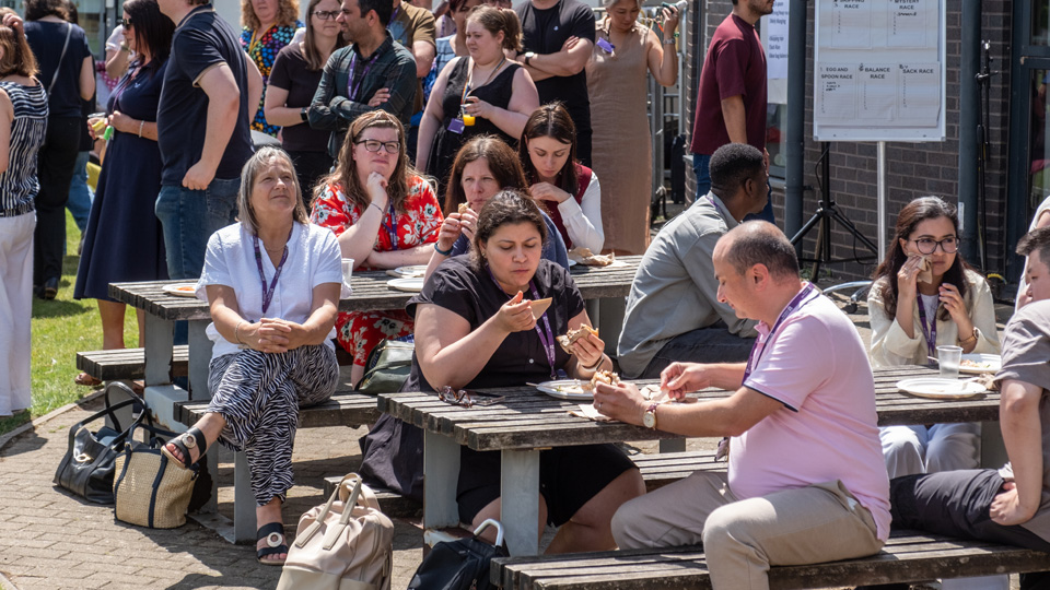 People gathered outdoors around picnic tables, eating, drinking, and talking near a brick building with posters